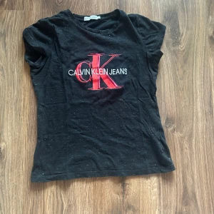 Svart t-shirt från Calvin Klein Jeans - Svart lite glittig t-shirt från Calvin Klein med stort rött och vitt logotryck på bröstet. Klassisk rund halsringning och korta ärmar. Den är av bra kvalitet eftersom den är äkta! 💓