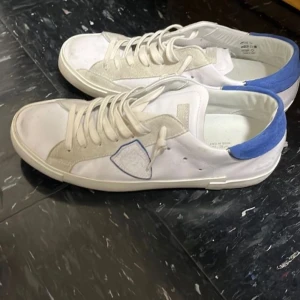 Vita sneakers med blå detalj - Snygga vita sneakers med beige detaljer och blå häl. Skorna har snörning och en dekorativ patch på sidan. Perfekta för en stilren och avslappnad look.