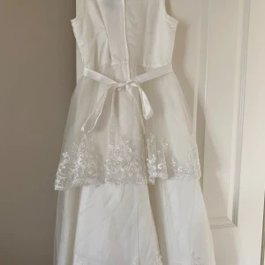 Vit långklänning med spets från Sessa Mode de Paris - Elegant vit långklänning med ärmlös design och vacker spetsdetalj upptill samt längs kjolen. Klänningen har ett tunt band i midjan och lager av tyll med broderade blommor. Perfekt för speciella tillfällen.