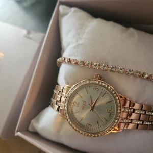 Guldig klocka med strass från Regal - Säljer en snygg guldig klocka från Regal med glittriga stenar runt urtavlan och på armbandet. Klockan har en klassisk rund form och matchas med ett armband med stenar. Perfekt accessoar för att lyfta din stil.