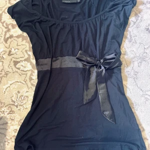 Svart t-shirt med rosett från KappAhl - Svart T-shirt från KappAhl med kort ärm och en dekorativ rosett i midjan. Klänningen har en markerad midja med ett brett band och är tillverkad i ett mjukt tyg.