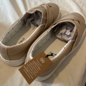 Beige slip-on sneakers med knutdetalj - Säljer ett par beige slip-on sneakers från Anna Field med snygg knutdetalj framtill. Skorna har vit platt sula och är tillverkade i textilmaterial. Perfekta för en avslappnad och trendig look.
