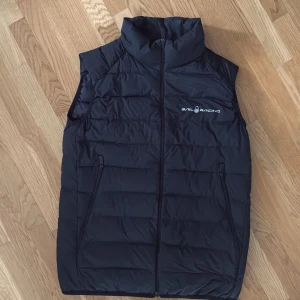 Svart dunväst från Sail Racing - Svart dunväst från Sail Racing med hög krage och dragkedja framtill. Använd ett fåtal gånger och är i bra skick. Västen har två fickor med dragkedja och logotyp på bröstet samt bak på nacken. Hör av er vid intresse! 