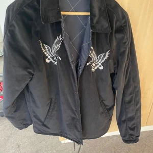 Svart jacka med broderade örnar - Snygg svart jacka med broderade örnar på bröstet och ryggen. Jackan har en klassisk krage och dragkedja framtill. Perfekt för en cool och unik stil.