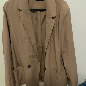Beige/brun kavaj från Gina Tricot - Säljer en stilren beige/brun kavaj från Gina Tricot. Kavajen har en klassisk design med två knappar framtill och fickor. 