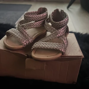 Flätade sandaler från Zara - Snygga flätade sandaler från Zara i en metallisk nyans. De har flera remmar som korsar över foten och en praktisk dragkedja bak för enkel på- och avtagning. Perfekta för sommarens alla äventyr!