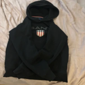 Svart hoodie från GANT - Säljer en svart hoodie från GANT med ett broderat emblem på bröstet. Tröjan har en klassisk design med huva och en stor ficka framtill. Perfekt för en avslappnad stil. Den är helt ny köpte den igår