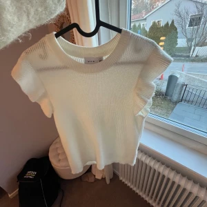 Vit stickad topp - Säljer en stilren vit ribbad topp från Vila. Toppen har korta ärmar med volangdetaljer och en rund halsringning. Perfekt för en chic och enkel look💖💖STORLEK:S❤️