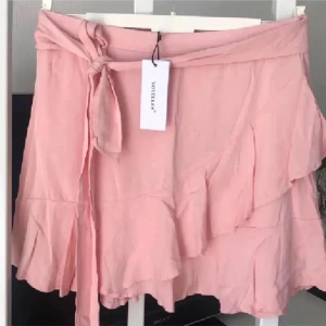 Rosa omlottkjol från Voyelles  - Söt rosa omlottkjol från Voyelles med volangdetaljer. Kjolen har en knytning i midjan och är perfekt för en somrig look. Den korta längden och det lätta materialet gör den idealisk för varma dagar. Storlek L på lappen men lite mindre i storleken . Midjemått 78 cm 