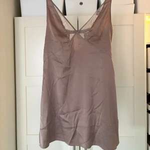 Beige klänning från H&M - Säljer en elegant beige/rosa klänning från H&M med spetsdetaljer och djup v-ringning. Klänningen är ärmlös och har en mjuk, glansig yta som ger en lyxig känsla. Perfekt för en stilren look.