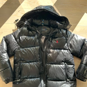 Svart pufferjacka från Polo Ralph Lauren - Snygg svart pufferjacka från Polo Ralph Lauren med huva och dragkedja. Jackan har en broderad logga på bröstet och flera praktiska fickor med dragkedjor. Perfekt för kyliga dagar. Skickar inom 24 timmar och pris kan diskuteras 