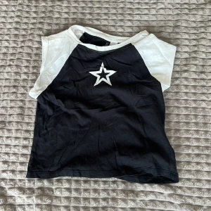 T-shirt - Säljer en cool svart och vit  topp med en stor vit stjärna på bröstet. Perfekt för en avslappnad stil. Toppen har en kontrasterande design med vita axlar och svart kropp.