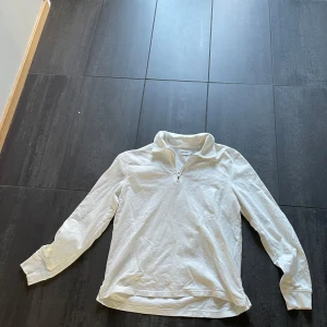 Vit tröja med dragkedja från East West - Säljer en stilren vit tröja från East West med dragkedja vid halsen. Tröjan har långa ärmar och en bekväm passform, perfekt för en avslappnad look.