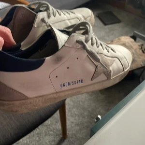 Vita sneakers från Golden Goose strlk 42 - Snygga vita sneakers från Golden Goose med en ikonisk stjärndetalj på sidan. Skorna har en blå häl och klassisk snörning. Perfekta för en stilren look. Dustbag och skolåda kommer med har tyvärr inget kvitto. Skorna är i extremt bra skick då de använts sparsamt 