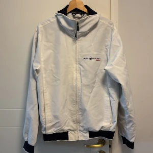 Vit jacka från Sail Racing - Säljer en stilren vit jacka från Sail Racing med dragkedja och broderad logga på bröstet. Jackan har långa ärmar och ribbade muddar i svart vid ärmslut och nederkant. Perfekt för en sportig look!