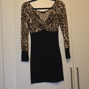 Leopardmönstrad klänning från Om Crazy - Snygg klänning med leopardmönstrad överdel och svart underdel. Klänningen har långa ärmar och en knutdetalj framtill. Perfekt för en djärv look!