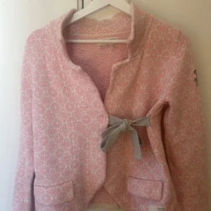Rosa mönstrad jacka med knytband från Odd Molly storlek 1 - Säljer en söt rosa jacka med ett diskret mönster och grått knytband framtill från Odd Molly. Jackan har långa ärmar och en broderad detalj på ärmen. Perfekt för en stilren look!
