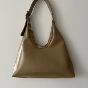 Beige axelväska Carin Wester med dragkedja - Snygg beige axelväska i syntetmaterial från C.W. Väskan har en stilren design med en praktisk dragkedja på sidan och justerbar axelrem. Perfekt för att bära dina dagliga nödvändigheter med stil.