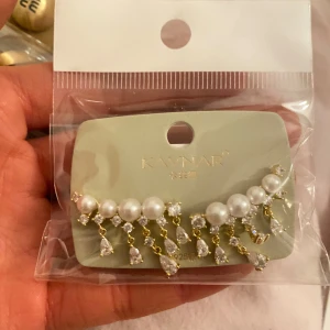 Örhängen med pärlor och kristaller från Kaynar - Eleganta örhängen från Kaynar med vita pärlor och gnistrande kristaller. Designen har en rad av pärlor med hängande kristaller i guldtonad infattning. Perfekt för att ge en lyxig touch till din outfit.