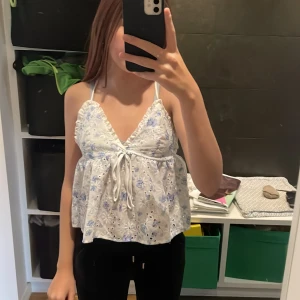 Vit blommig linnetopp - Säljer en söt vit linnetopp med blå blommor och broderade detaljer. Toppen har en v-ringning och knytband framtill. Perfekt för sommardagar!