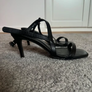 Svarta klackskor - Snygga svarta sandaler från Tiamo med smala remmar och en elegant klack. Perfekta för en stilren look. Remmarna ger en säker passform och den fyrkantiga tån ger en modern touch.