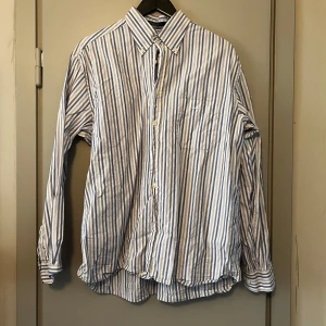 Randig skjorta från GANT - Säljer en stilren randig skjorta från GANT i blått och vitt. Skjortan är tillverkad av egyptisk bomull och har en normal passform. Den har långa ärmar och knappar framtill samt en bröstficka med broderad logotyp.