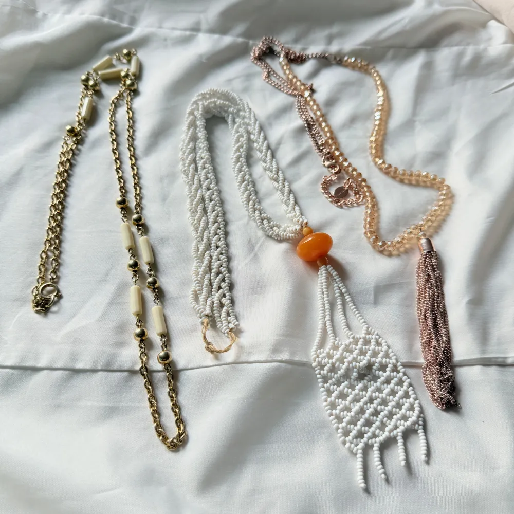 En samling av unika halsband: ett guld- och vitfärgat halsband med kedjedetaljer, ett vitt pärlhalsband med orange detalj, och ett rosa halsband med tofs. Perfekt för att mixa och matcha till olika outfits.. Asusteet.