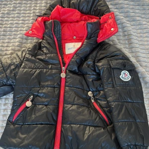 Svart jacka - Snygg svart dunjacka från Moncler med röd insida och huva. Jackan har dragkedja och knappar samt praktiska fickor med dragkedjor. Perfekt för kyliga dagar.