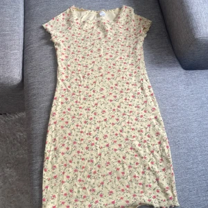 Blommig klänning i bomull - Söt blommig klänning i ljusgul bomull med små rosa och gröna blommor. Klänningen har korta ärmar och en bekväm passform. Perfekt för varma dagar!