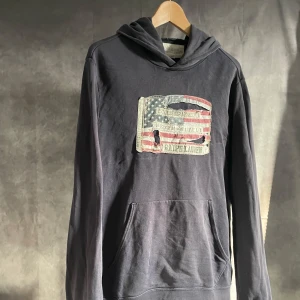 Ralph Lauren hoodie  - Säljer en mörkblå hoodie från Ralph Lauren med ett vintageinspirerat tryck av en amerikansk flagga på framsidan. Tröjan har en klassisk känguruficka och långa ärmar. Perfekt för en avslappnad stil.