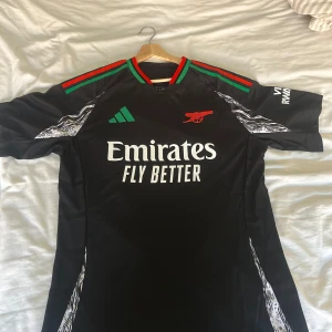 Svart Arsenal  fotbollströja från Adidas - Säljer en svart fotbollströja från Adidas med röda och gröna detaljer på axlarna. Tröjan har korta ärmar och fina svartvita mönster  lite här och var. Tröjan är använd i några månader men har tyvärr blivit för liten för mig därför säljer jag denna. Storleken är M men passar även S. Kontakta mig vid fler frågor!
