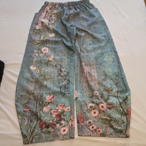 Blommiga byxor med elastisk midja - Snygga byxor med ett vackert blommönster i rosa och gröna nyanser. De har en elastisk midja för extra komfort och en lös passform. Perfekta för en avslappnad stil med en touch av elegans.