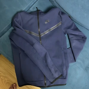 Blå hoodie från Nike - Säljer en blå hoodie från Nike med dragkedja och huva. Jackan har en diskret logga på bröstet och en stilren design med svarta detaljer. Perfekt för en sportig look.