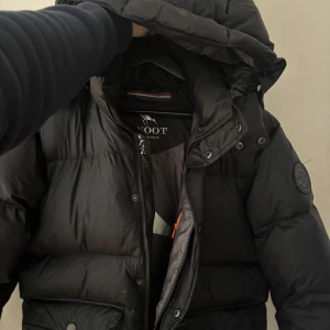 Svart Snoot jacka - Säljer en svart pufferjacka från Snoot med huva och flera praktiska fickor. Jackan har en stilren design med knappar och dragkedja framtill. Perfekt för kyliga dagar. Använd i en säsong, ordniare pris 6000kr