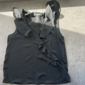 Svart ärmlös blus med volangdetaljer från Zara - Snygg svart ärmlös blus från Zara med volangdetaljer längs halsringningen. Perfekt för en stilren look. Säljer pga för liten - sparsamt använt! Passar både till jeans och kostym byxor