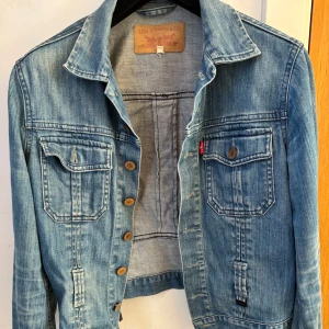 Blå jeansjacka från Levi's - Snygg blå jeansjacka från Levi's med knappar framtill och två bröstfickor. Jackan har en klassisk krage och snörning i ryggen för en unik touch. Perfekt för en avslappnad stil.