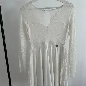 Elegant vit klänning med vacker spetsdetalj och lång ärm. Klänningen har en v-ringad design och en mjuk, flödande kjol. Perfekt för speciella tillfällen. Den har även dragkedja på baksidan. 