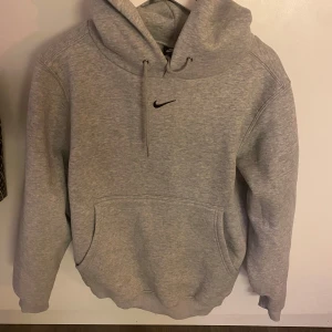Grå hoodie från Nike - Säljer en klassisk grå hoodie från Nike med en liten svart logga på bröstet. Tröjan har en stor ficka framtill och en justerbar huva. Perfekt för en avslappnad stil.
