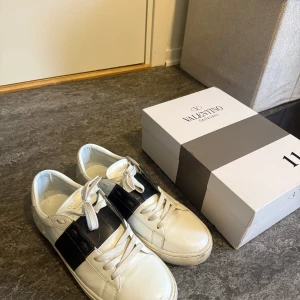  Valentino Garavani  - Säljer ett par snygga vita Valentino Garavani sneakers med bred svart rem över sidan. Skorna har rund tå, vita snören och diskret Valentino-logga på plösen. Perfekta för dig som gillar stilrena och exklusiva sneakers. Box ingår!