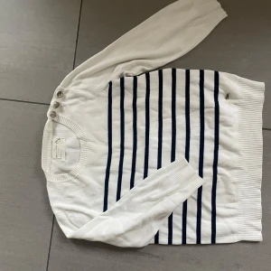 Vit och marinblå randig tröja från Holly & Whyte - Vit långärmad tröja från Holly & Whyte med marinblå horisontella ränder och tre knappar på axeln. Tröjan har ribbade muddar och är gjord i mjuk bomull. Perfekt för en klassisk och stilren look.