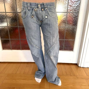 Unika jeans med broderad text  - Säljer ett par blå jeansbyxor med coola detaljer som nitar, broderad text på bakfickan och flera knappar framtill. Byxorna har en rak passform och markerade sömmar som ger en unik look. Perfekta för dig som vill sticka ut.