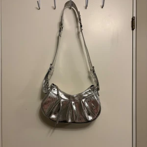 Silver axelväska från Gina Tricot - Snygg axelväska i metallic silver från Gina Tricot. Väskan har en bred justerbar axelrem med nitar och detaljer, samt veckad framsida. Perfekt för att addera lite extra bling till din outfit.