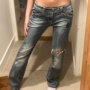 Lågmidjade y2k jeans - Säljer ett par blå jeans med slitningar, patch på låret och snygga broderade detaljer på bakfickorna. Innerbenslängden är 74cm och midjan är 38cm rakt över💕(reserverad!!)