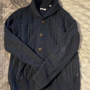 Mörkblå stickad kofta från Jack & Jones - Säljer en mörkblå stickad kofta från Jack & Jones med grovt kabelstickat mönster och stora knappar framtill. Koftan har hög krage och ribbade muddar vid ärmslut.