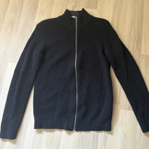 Mörk blå stickad kofta från Selected Homme - Säljer en mörkblå stickad kofta från Selected Homme som är stickad med hög krage och dragkedja framtill. Tröjan har lång ärm och ribbade muddar vid ärmslut och nederkant. Perfekt för lager-på-lager och enkel att matcha. Koftan är i storlek M och är använd en gång 