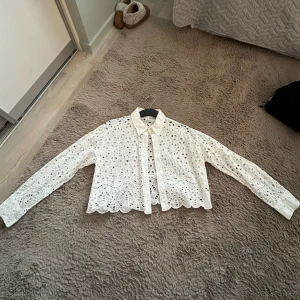 Vit broderad blus från H&M - Superfin vit blus från H&M med broderade detaljer och hålmönster. Blusen har långa ärmar, knappar framtill och en croppad passform. Perfekt för vår och sommar när du vill ha något luftigt och snyggt.