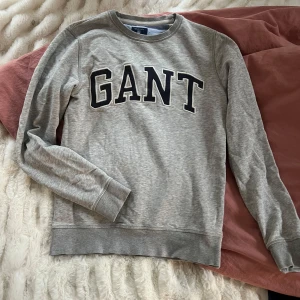 Grå sweatshirt från GANT - Säljer en klassisk grå sweatshirt från GANT med svart och vit logotyp framtill. Tröjan har rund halsringning och långa ärmar. Perfekt för en avslappnad och stilren look.
