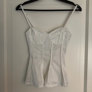Vit korsett-topp med smala axelband - Säljer en snygg vit korsett-topp med smala ställbara axelband och markerade sömmar. Toppen har en figurnära passform och lätt utsvängd nederdel. Längd framtill 32cm