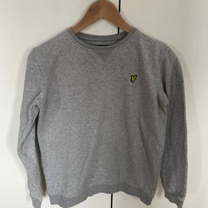 Grå sweatshirt från Lyle & Scott  - Klassisk grå sweatshirt från Lyle & Scott med rund halsringning och broderad gul logga på bröstet. Tröjan är varsamt använd och inga defekter. Storleken är 12/13 år vilket motsvarar ungefär 164. Tröjan har långa ärmar och ribbade muddar vid ärmslut och nederkant. Perfekt för en avslappnad stil men som även är stilren.