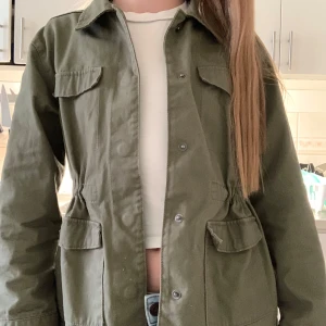 Grön jeansjacka från Kappahl - Säljer en olivgrön jacka från Kappahl i storlek 146. Jackan har klassisk krage, tryckknappar framtill, två stora fickor och markerad midja med resår. Perfekt för lager-på-lager och enkel att matcha med olika outfits. 💚